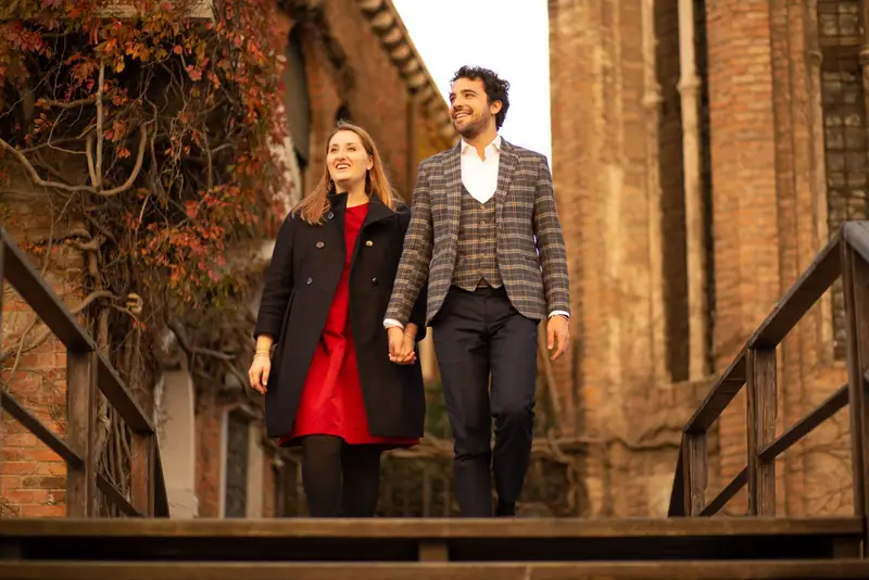 Michela e Massimiliano camminano mano nella mano su una passerella di legno, fra muri di mattoni e foglie rosse