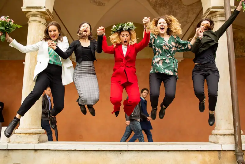 Cinque amiche saltano insieme sotto un portico per festeggiare una laurea, una con la corona d’alloro