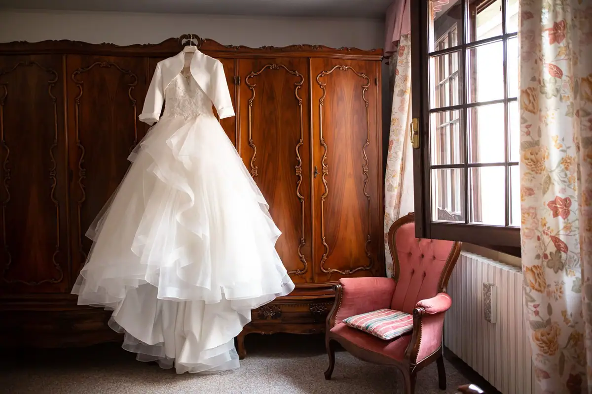 L’abito da sposa appeso a un armadio di legno, accanto alla finestra, in una camera con la poltroncina rosa
