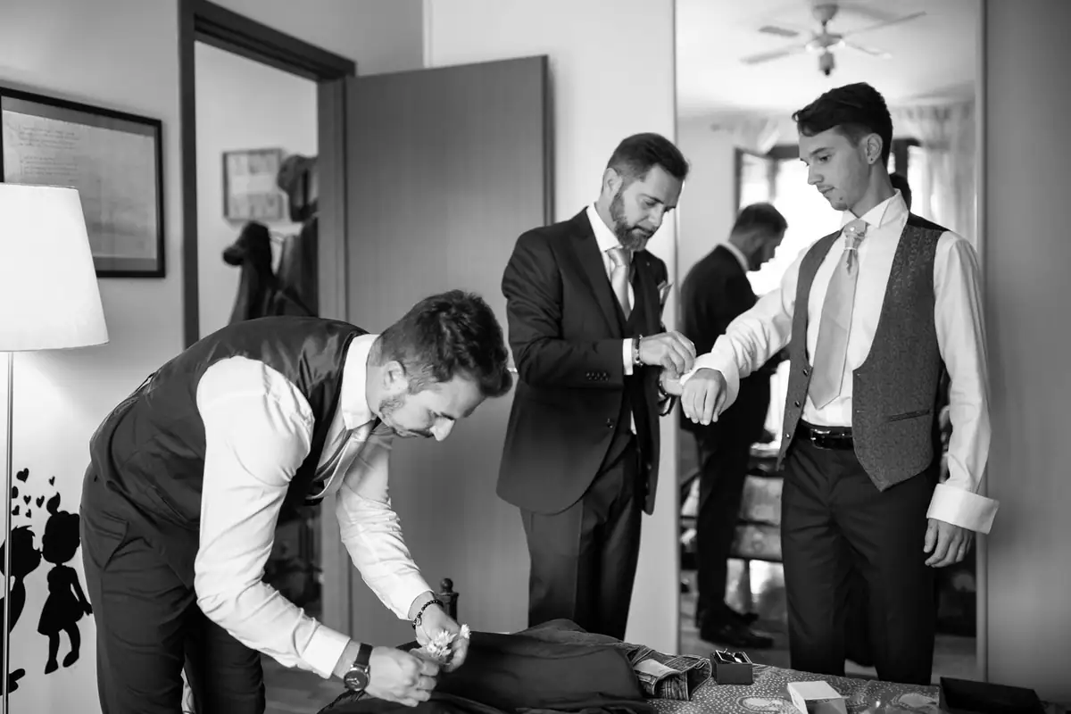 In bianco e nero, lo sposo si sistema la giacca mentre intorno a lui ci si prepara