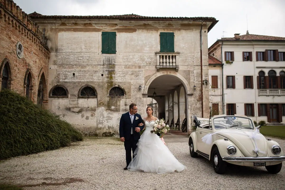 La sposa e chi l’accompagna camminano sulla ghiaia davanti alla facciata antica della villa, il maggiolino accanto