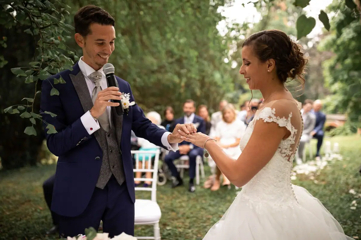 Lo sposo legge le promesse al microfono, la sposa lo guarda e ride, fra gli alberi del parco