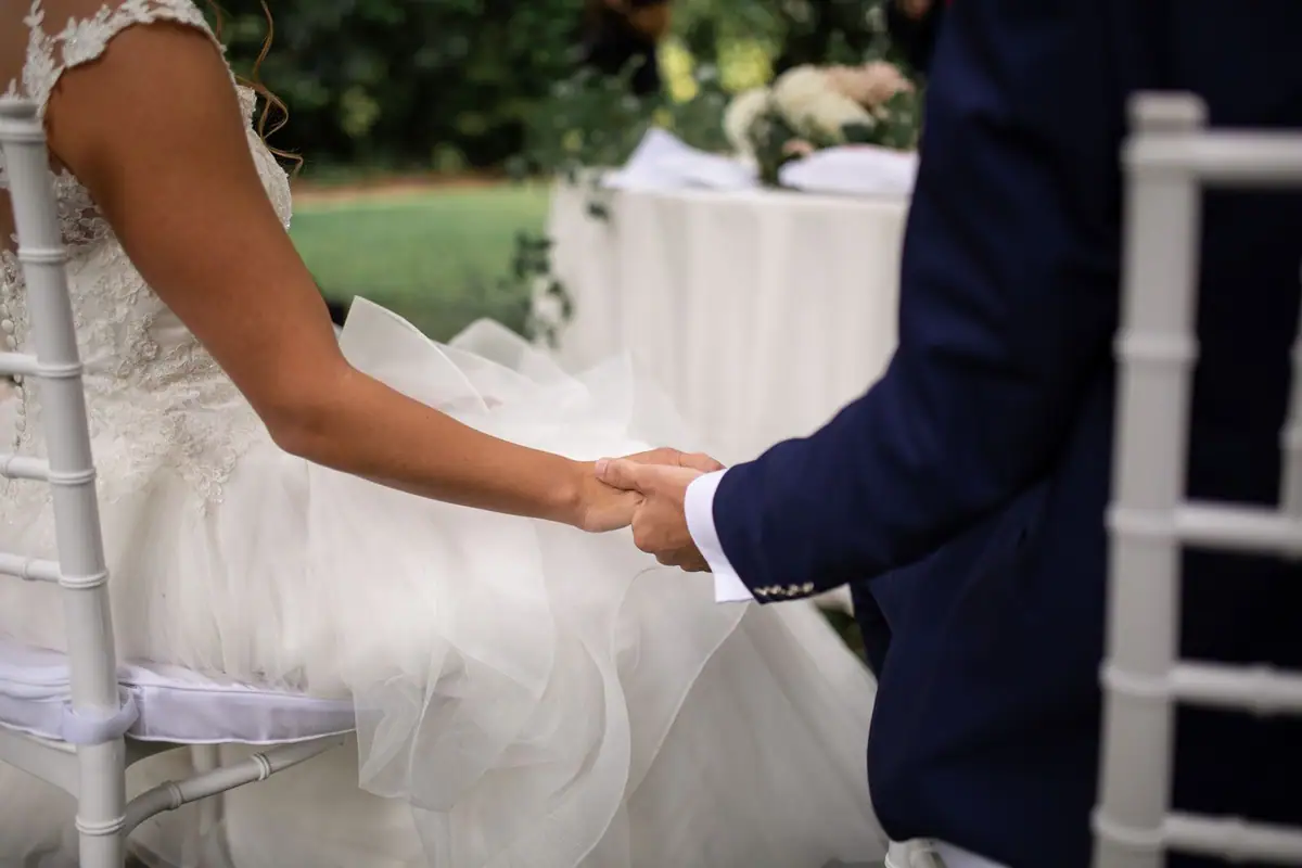 Le mani degli sposi si stringono sopra il tulle dell’abito, sulle sedie bianche