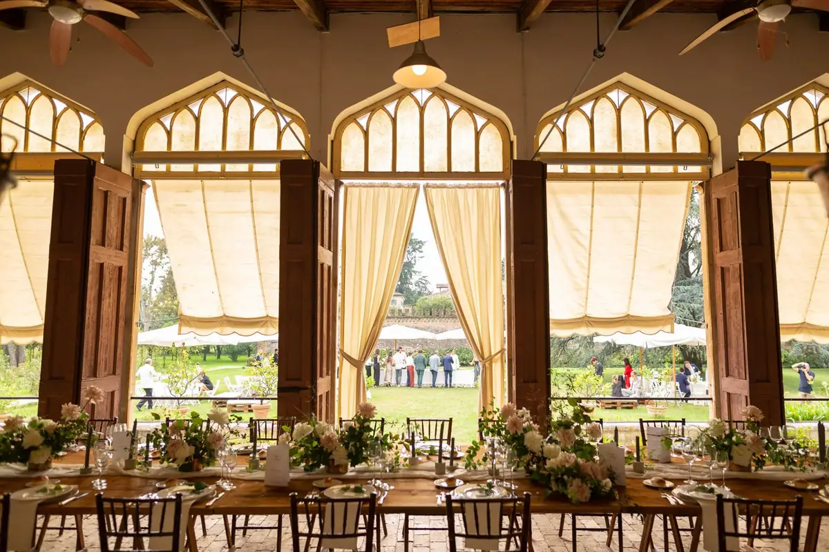 L’aranciera apparecchiata: tavoli lunghi di legno, fiori bianchi, le grandi finestre ad arco aperte sul giardino