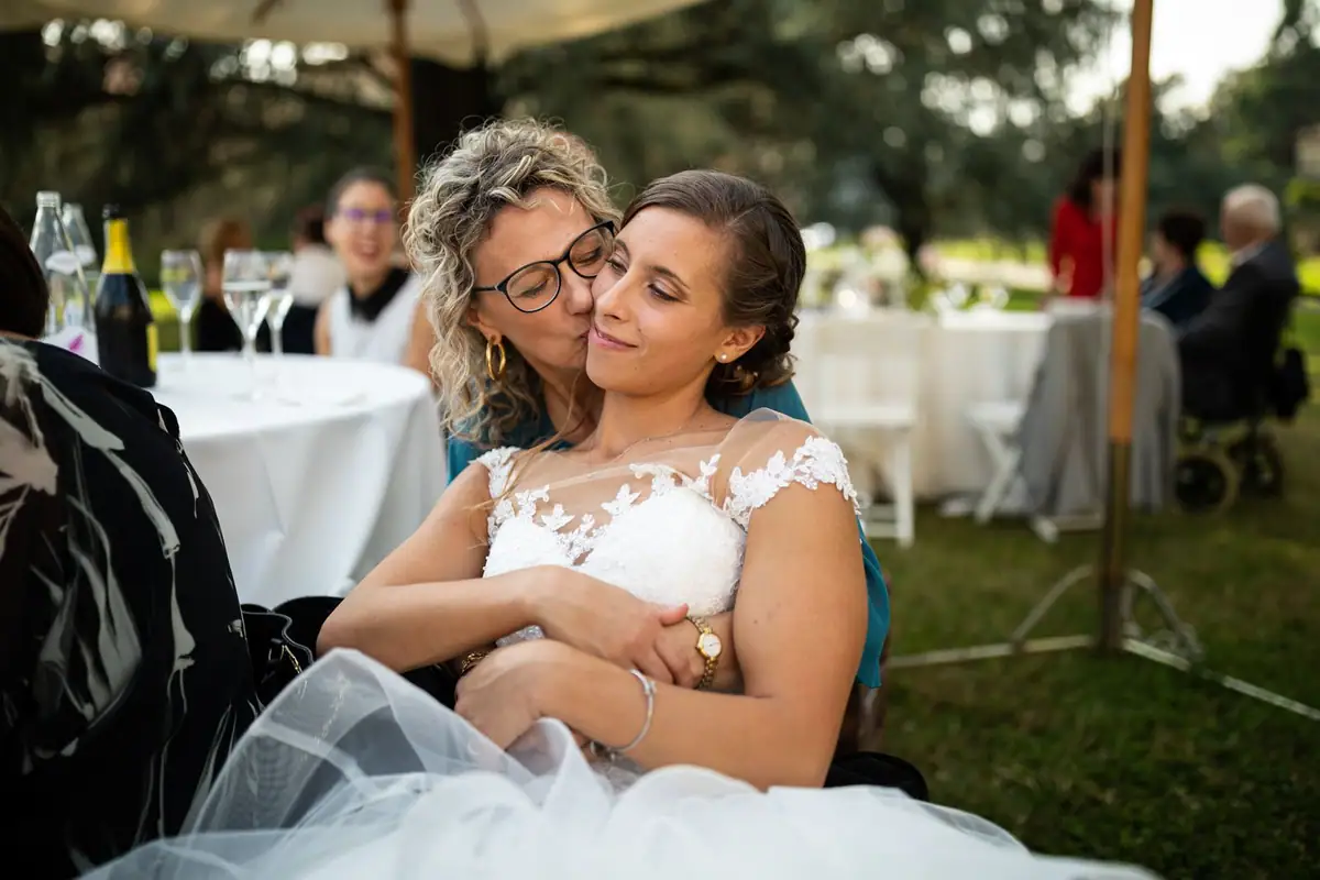 Un’invitata bacia la sposa sulla guancia, al tavolo, gli occhi chiusi